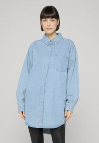 Woman with short dark hair wearing an oversized light blue denim shirt and black pants against a plain gray background.