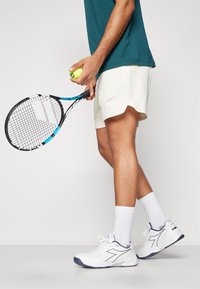 Weiße Tennisschuhe mit marineblauen Akzenten, weiße Socken tragend. Hellbeige Sportshorts, hält einen Tennisschläger und einen grünen Tennisball.
