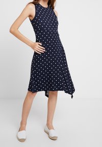 Námořnické bezrukávové šaty s bílými puntíky, přiléhavý korzet, rozšířená sukně a asymetrický lem, doplněné bílými espadrilami.