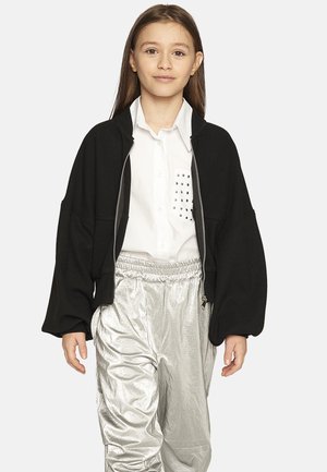 Cardigan nero con zip e maniche oversize, indossato sopra una camicia bianca con dettagli di tasca borchiati, abbinato a pantaloni metallizzati argento.