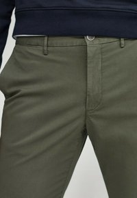 Primo piano di una persona che indossa pantaloni verde oliva con bottone e zip frontali, abbinati a un maglione blu scuro.