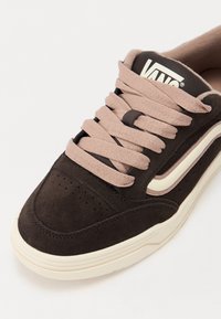 Brown suede sneaker with light pink laces, cream accents, and perforated detailing on the toe cap. Logo on the tongue. Flat sole design.
