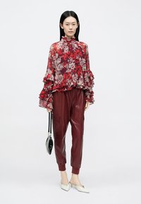 Camicia trasparente rossa con stampa floreale e volant, abbinata a pantaloni jogger in pelle bordeaux profondo. Accessori includono una borsa nera e ballerine crema con punta appuntita.