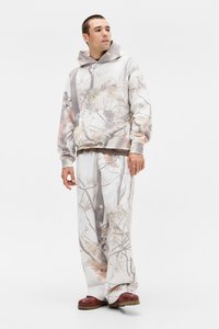 Conjunto de sudadera con capucha y pantalones de camuflaje en colores claros con patrones de hojas, ajuste holgado y mangas largas. Calzado marrón completa el atuendo.