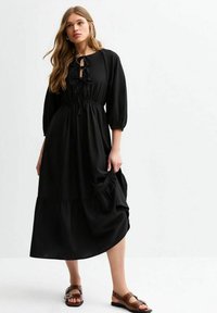 Black midi dress with a round neckline, tied front detail, and elastic waist. Features puff sleeves and a tiered hem. Worn with brown sandals.