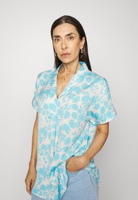 Blusa de manga curta floral azul clara com colarinho, apresentando um corte descontraído e tecido suave. Contraste de cor notável com fundo branco.