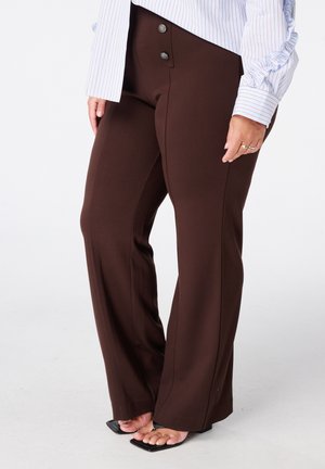 Vrouw draagt donkerbruine wijde broek met twee knopen en zwarte sandalen met vierkante neus, en een wit gestreepte blouse met ruches aan de mouwen.