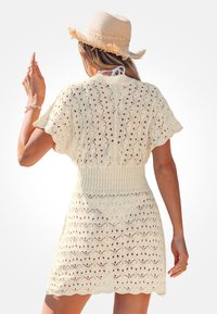 Mujer con un vestido de ganchillo color crema y un sombrero de paja, de pie de espaldas a la cámara y con una mano levantada.
