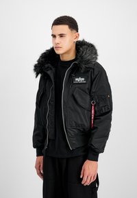 Schwarz gepolsterte Jacke mit Kapuze aus Kunstfell, Reißverschluss und Armtasche. Merkmale ein Logodesign und ein kontrastierendes rotes Etikett. Strukturierte Gestaltung.