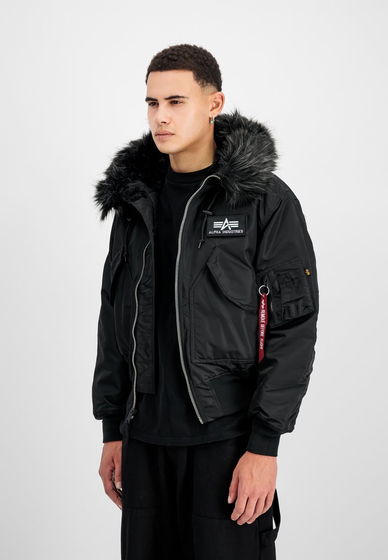 Schwarz gepolsterte Jacke mit Kapuze aus Kunstfell, Reißverschluss und Armtasche. Merkmale ein Logodesign und ein kontrastierendes rotes Etikett. Strukturierte Gestaltung.