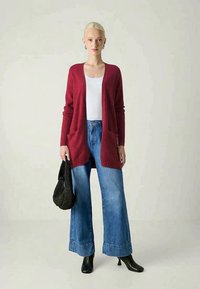 Cardigan de malha vermelha com bolsos, blusa branca, jeans de perna larga azul, botas pretas de tornozelo e uma bolsa preta texturizada.