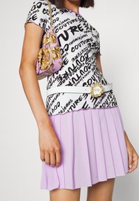 Woman wearing a white and black graphic Versace Jeans Couture top, lavender pleated skirt, white belt with gold buckle, and a purple patterned handbag.