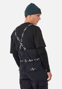 Schwarzes T-Shirt mit weißen Logo-Trägern, die sich auf dem Rücken kreuzen, kombiniert mit dunklen Hosen und einer gerippten dunkelblauen Mütze.