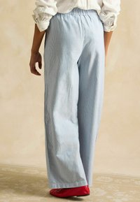 Svetlomodré pruhované nohavice s širokými nohavicami a elastickým pásom, hladký povrch a v popredí je vidno červenú plochú obuv.
