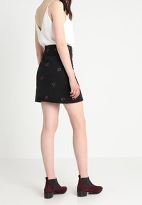 Kvinna i vit ärmlös topp, svart blommig minikjol och vingård färgade ankelkängor, stående mot en enkel vit bakgrund.