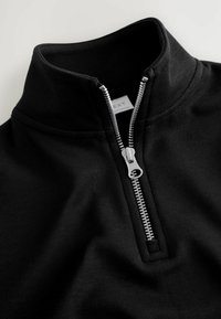 Sort pullover med høj krave, med en sølvfarvet lynlås og ribbet tekstur. Minimalistisk design, mærket med brandet "NEXT."