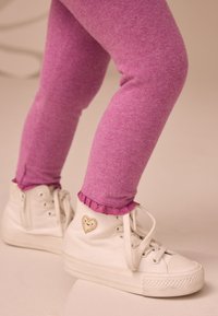 Valged kõrge säärega tennised, millel on paelad ja kuldse südame tikand, koos roosade leggingsidega, millel on volangidest ääris.