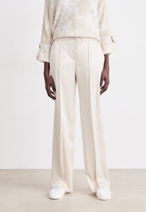 Pantalons larges beige clair avec une finition texturée, dotés d'une taille à cordon et de plis sur les côtés, associés à des baskets blanches.