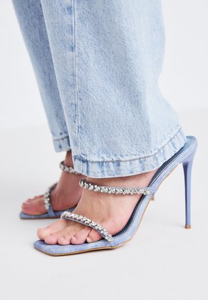 Sandálias de salto alto em denim azul claro, com duas tiras adornadas com cristais e um salto fino. Jeans casuais cobrem parcialmente o calçado.