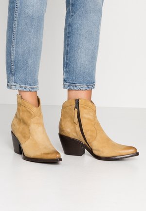 Bottines en daim camel avec fermeture éclair latérale et talons blocs portées avec un jean court bleu clair sur fond uni.