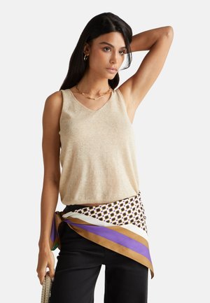 Beige mouwloze top in zachte stof met een V-hals; gecombineerd met een patroonwrap in witte, bruine en paarse tinten. Zwarte broek.