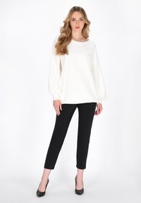 Witte oversized trui met ballonmouwen, gecombineerd met zwarte slim-fit broek en zwarte hoge hakken, tegen een neutrale achtergrond.