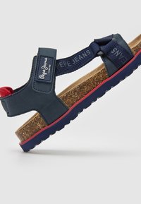 Tamsiai mėlynas sandalai su kamštiniu padu, tekstūruotos dirželiai mėlynos ir raudonos spalvų, su prekės ženklo etikete. Pakėlimas iš guminiu padu su griovelių raštu.