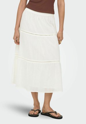 Femme portant une longue jupe blanche à volants avec bordure en dentelle, un top marron sans manches et des tongs noires avec semelles à motifs.