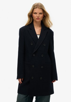 Marineblauwe dubbelzijdige wollen jas met grote zwarte knopen, pieklapels en twee voorzakken, gecombineerd met een lichte top en losse denim jeans.