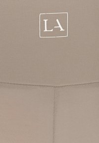 Beige ribbed sporttop met een klein wit "LA" logo gecentreerd binnen een rechthoekige omlijning. Gladde textuur met rekbare stof.