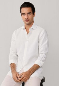 Homme aux cheveux foncés portant une chemise blanche boutonnée et un pantalon clair, assis sur un tabouret noir devant un fond gris uni.