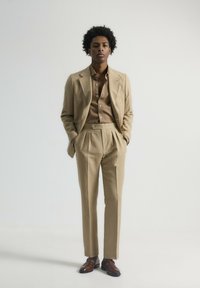 Traje de lino beige con chaqueta ajustada y pantalones con pliegues. Camisa de botón marrón a juego por debajo. Zapatos de vestir de cuero marrón, diseño minimalista.