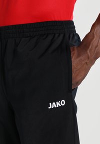 Pantaloni sportivi neri realizzati in materiale liscio, con una vita elastica e tasca laterale. Logo "JAKO" ricamato in bianco sulla gamba.