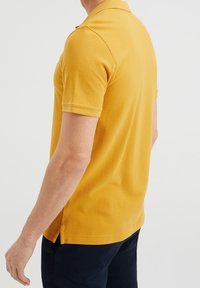 Polo t-shirt i senapsgul färg tillverkad av strukturerad tyg, med klassisk krage, korta ärmar och sidoflikar för ökad rörelsefrihet.