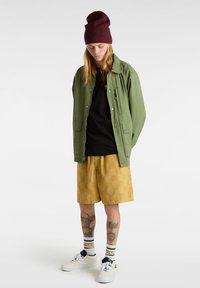 Zielona lekka kurtka, czarna koszulka, żółte szorty w wzory, białe sneakersy oraz bordowa czapka i paski na skarpetkach, z tatuażami na nogach.