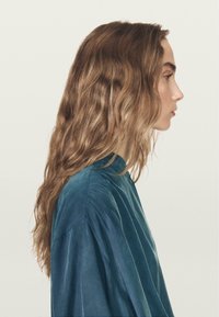 Profil af en person med langt, bølget, brunt hår, iført en tealfarvet, flydende top med blød tekstur og en høj halsudskæring.