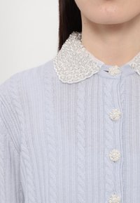 Gros plan sur un pull en maille torsadée bleu clair avec col orné de perles et cristaux, et boutons décoratifs assortis.