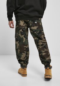 Camo cargo pants in dark green, black, and brown, featuring side pockets, adjustable cuffs, and a fabric texture. Brown boots visible below.