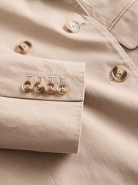 Veste ajustée beige en tissu avec une texture lisse. Présente deux grands boutons et cinq petits boutons au niveau des poignets, avec des détails de broderie décoratifs.