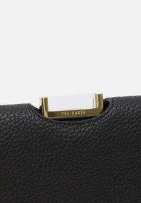 Borsa in pelle nera con texture, dotata di chiusura dorata con la scritta "Ted Baker" incisa sul bordo metallico.