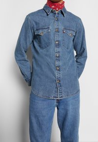 Chemise en jean bleu moyen à manches longues, fermeture par boutons, deux poches poitrine, et ourlet arrondi. Portée avec un bandana rouge en accent.