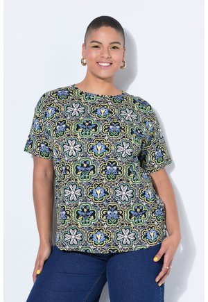 Lächende Person mit kurzen Haaren trägt goldene Creolen, gelbes Augen-Make-up, ein gemustertes Blumenhemd und dunkelblaue Jeans vor einem weißen Hintergrund.