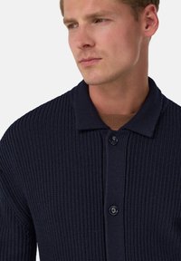 Mörkblå ribbad cardigan med krage, utrustad med två knappar och en texturerad stickad design. Plagget har ett ljusbrunt innerfoder som är synligt.