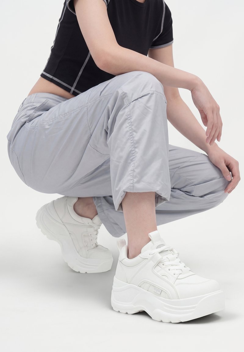 Personne accroupie portant un pantalon gris clair, un haut noir à manches courtes et des baskets compensées blanches épaisses sur fond blanc.