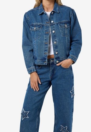 Vrouw draagt een blauwe spijkerjas en bijpassende jeans met versleten sterretjes, wit cropped top zichtbaar onder de jas.