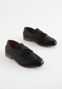 Mocassins noirs en cuir avec un panneau en maille texturée, un gland décoratif et un petit talon en bois. Présente des détails cousus sur les côtés.