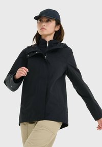 Veste imperméable noire avec capuche, fermeture éclair et poches latérales ; portée par-dessus un pull polaire noir à fermeture éclair. Un pantalon de couleur neutre complète la tenue.