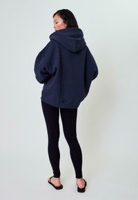 Felpa blu scuro oversize con un grande cappuccio, polsini e orlo a coste. Abbinata a leggins neri e infradito nere, su uno sfondo chiaro.