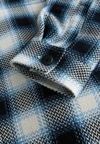 Modrá a čierna károvaná flanelová látka so zloženým lemom, vybavená čiernym gombíkom a štruktúrovaným tkaním. Vzor zahŕňa štvorce a rybiu kosť.