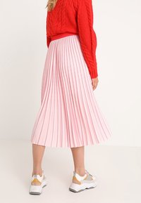 Jupe midi plissée rose avec une surface texturée, accompagnée d'un pull en maille rouge et de baskets blanches avec des accents gris et jaunes.